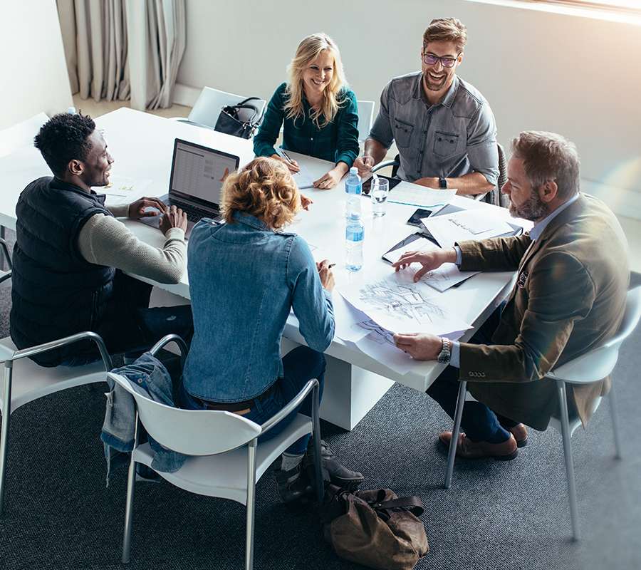 A group of people sit around a meeting table with their internet devices smiling and discussing business.