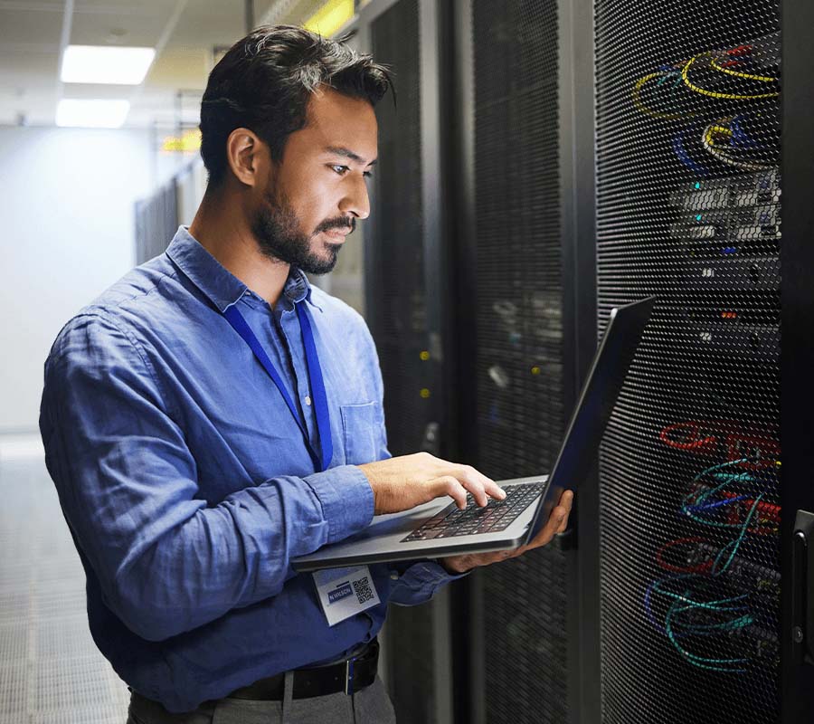 A network technician holding a laptop stands next to servers.