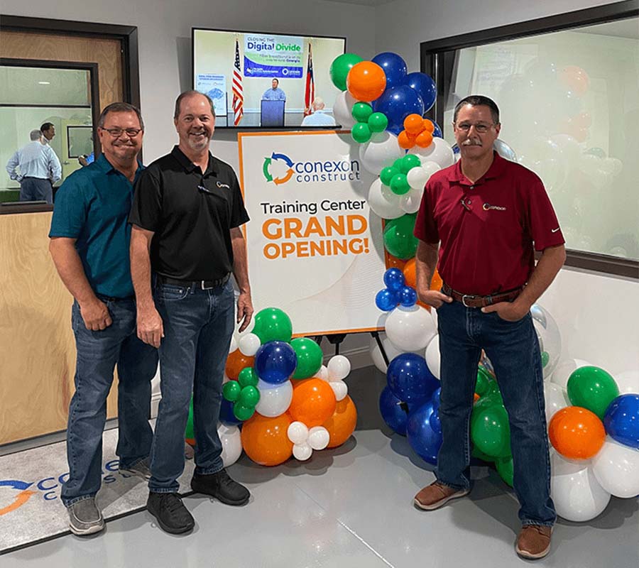 Three men smile and stand next to a display poster during the grand opening of Conexon Construct training center.